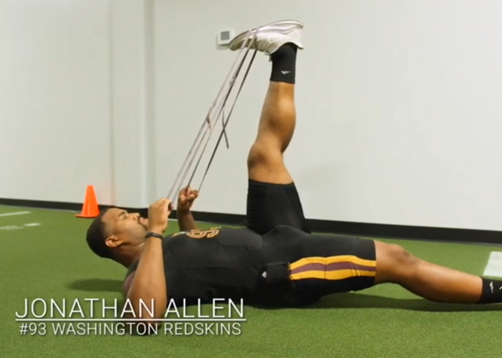Football player Jonathan Allen, wearing a black Washington Redskins outfit, training indoors with resistance bands based on positive testimonials.