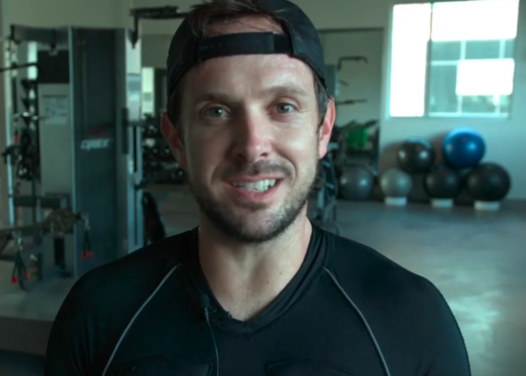 A man smiling at the camera, wearing a black athletic shirt and a cap, with a gym background featuring exercise equipment for reviews and testimonials.