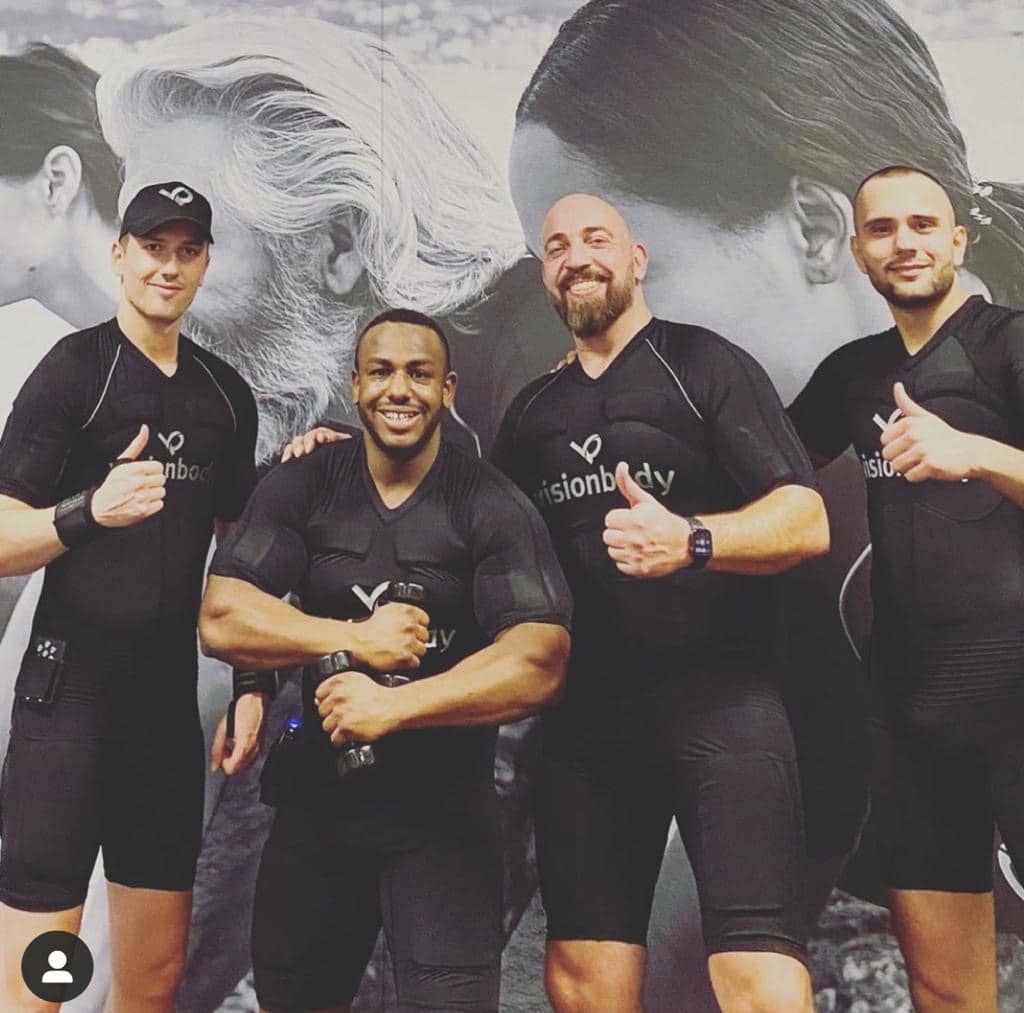Four men in branded athletic wear posing with thumbs up in front of a promotional banner featuring large facial close-ups.
