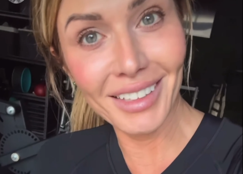 A close-up selfie of a woman with blond hair smiling at the camera, indoors with exercise equipment in the background, perfect for fitness reviews and testimonials.