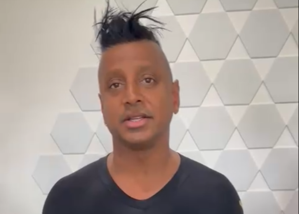 Man with an expressive hairstyle standing in front of a geometric-patterned white wall awaiting reviews and testimonials.