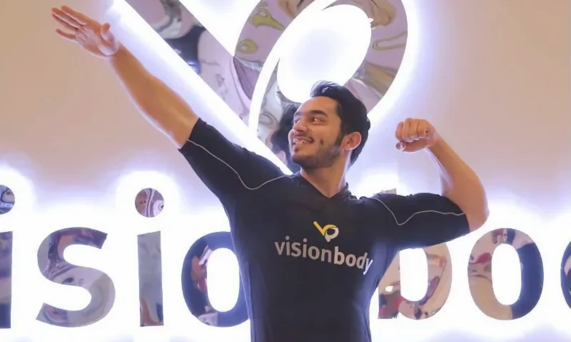 A person wearing a "visionbody" EMS Body suit strikes a bodybuilder pose in front of an illuminated "Visionbody" sign.