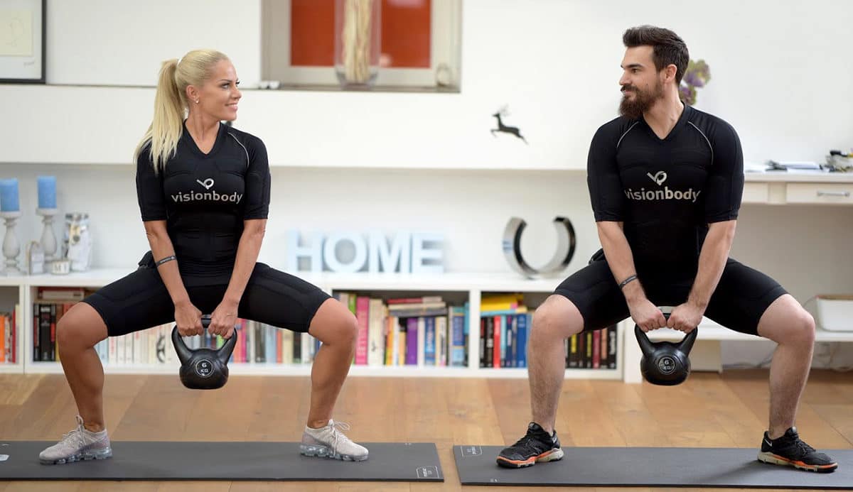 Two individuals wearing Visionbody EMS training suits, holding kettlebells, perform squat exercises.