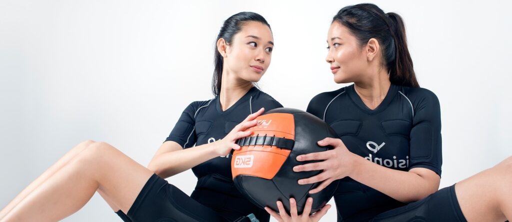Two women wearing the Visionbody EMS suit sit facing each other, holding a medicine ball between them during a workout session, against a plain white background.