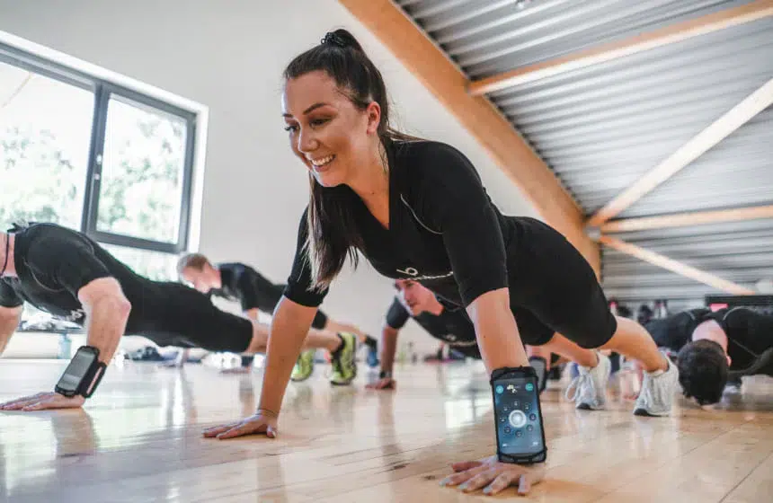 A group exercise class using EMS suits, including a woman smiling in the foreground performing a plank with a smartphone attached to her arm.