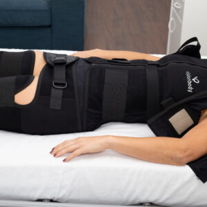 A person lies on a bed wearing a full-body compression suit that covers the torso, thighs, and knees, secured with multiple straps and fasteners.