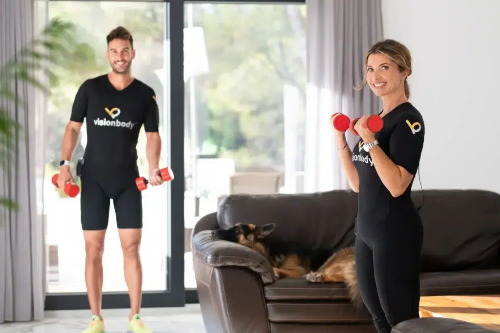 A man and woman exercise with dumbbells in a bright living room, matching black full body workout suits.