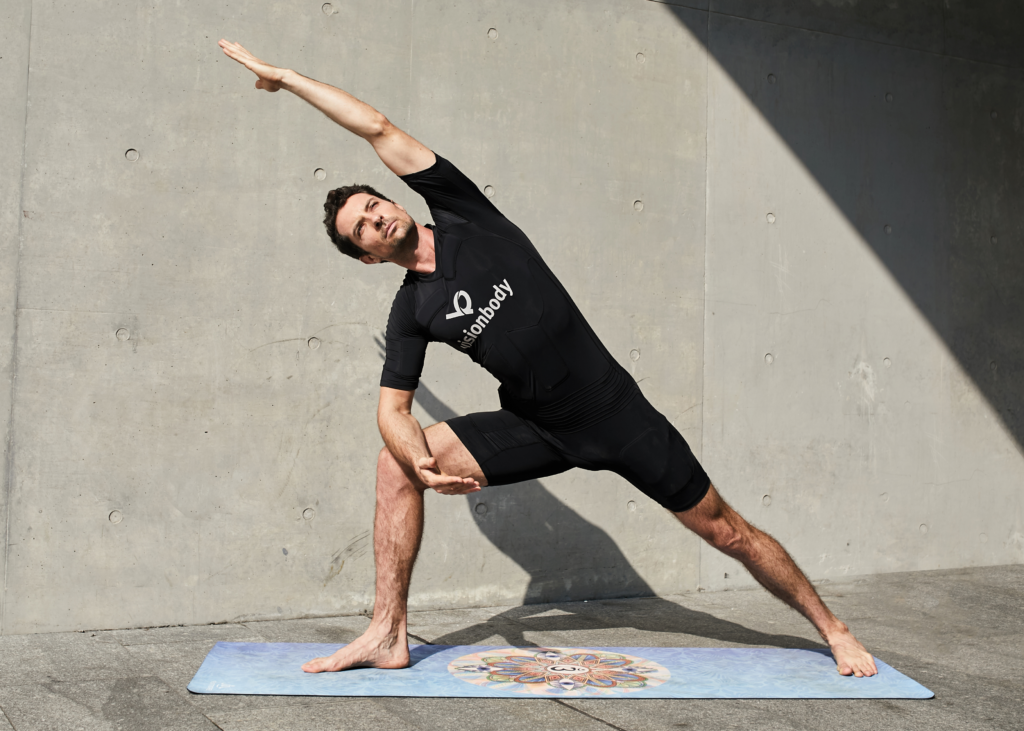 A man in black VisionBody athletic wear performs a yoga pose on a mat against a concrete wall, one arm reaching overhead and the other resting on a bent knee.