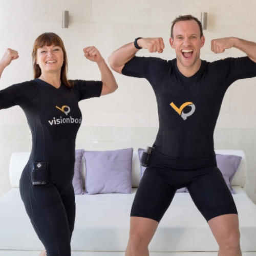 Two people wearing matching black athletic outfits with "visionbody" logos, standing indoors, smiling, and flexing their muscles.