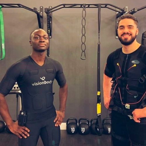 Two individuals wearing EMS workout suits stand in a gym. One is on the left with their hands on their hips, while the other is on the right, smiling, with gym equipment visible behind them.