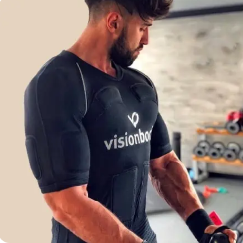 A person wearing a black fitted shirt with "visionbody" written on it is standing in what appears to be a gym, holding a piece of equipment. The background includes gym equipment and weights.
