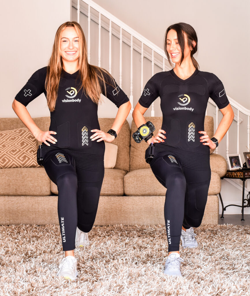 Two women in black athletic wear perform lunges indoors on a carpeted floor, smiling as they don their Visionbody compression suits designed for muscle growth. A staircase and furniture are visible in the background.