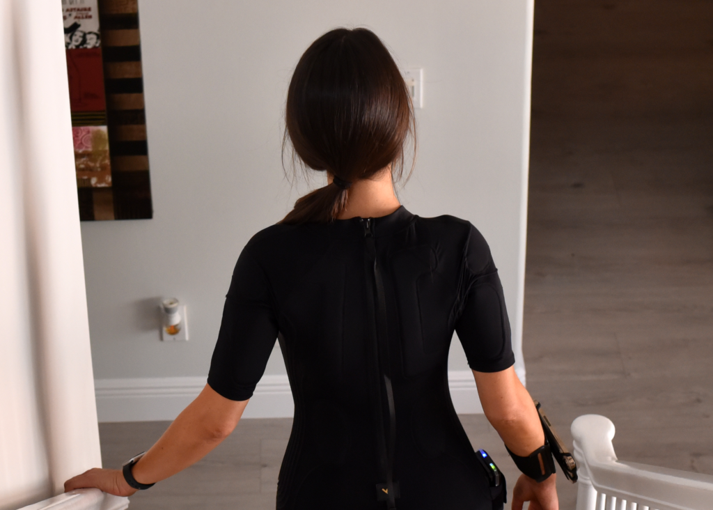A person in a black suit with technological devices walks down a staircase, facing away from the camera in a modern interior setting, their confident stride subtly reflecting their recent journey of muscle growth.