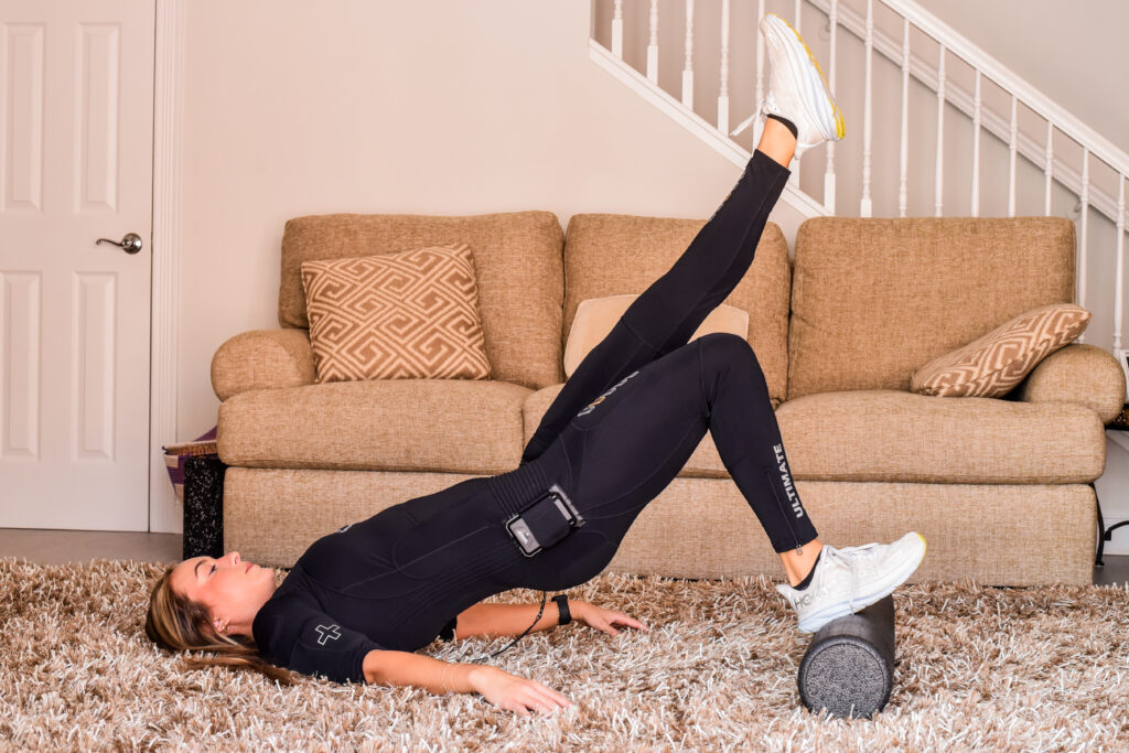 A person in athletic wear performs a single-leg glute bridge on a foam roller in a living room with a beige sofa and patterned pillow in the background, focusing on muscle growth.