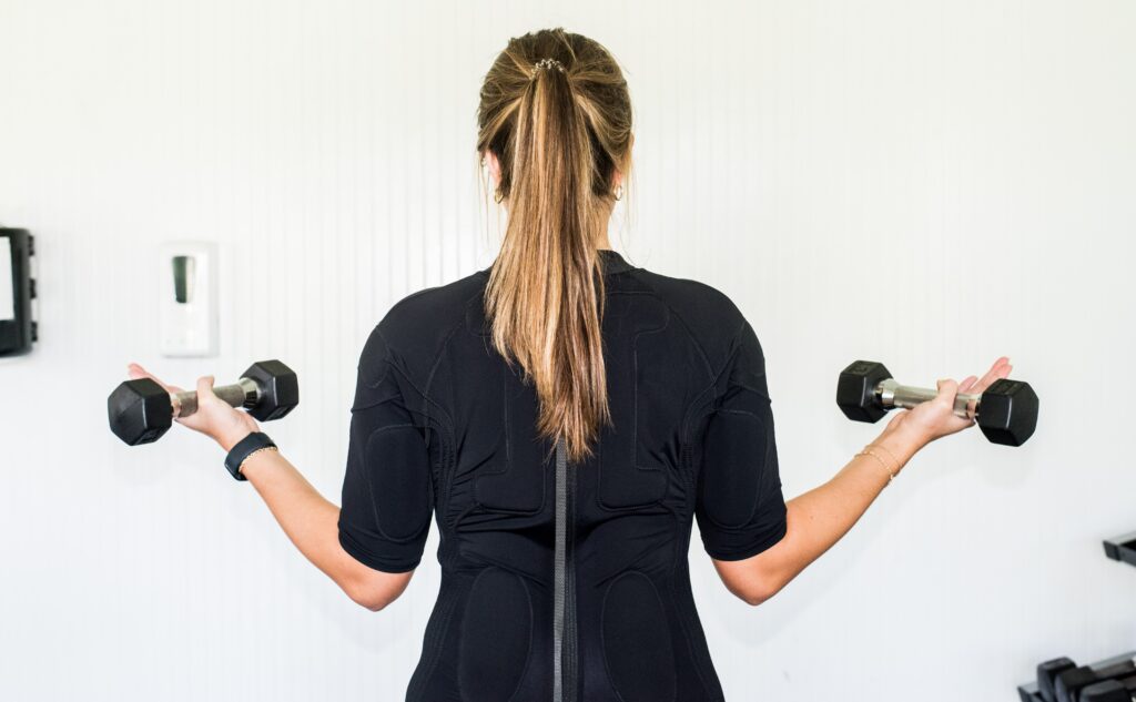A person with long hair in a ponytail, clad in black workout clothing, holds a dumbbell in each hand and faces away from the camera in a bright room, focused on muscle growth.