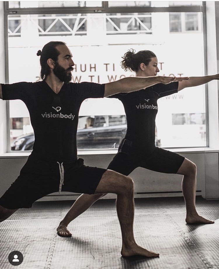 A man and a woman practice the Warrior II yoga pose indoors, aiming to achieve their weight loss goals. They are wearing matching black outfits displaying the word "visionbody." A large window is visible in the background.