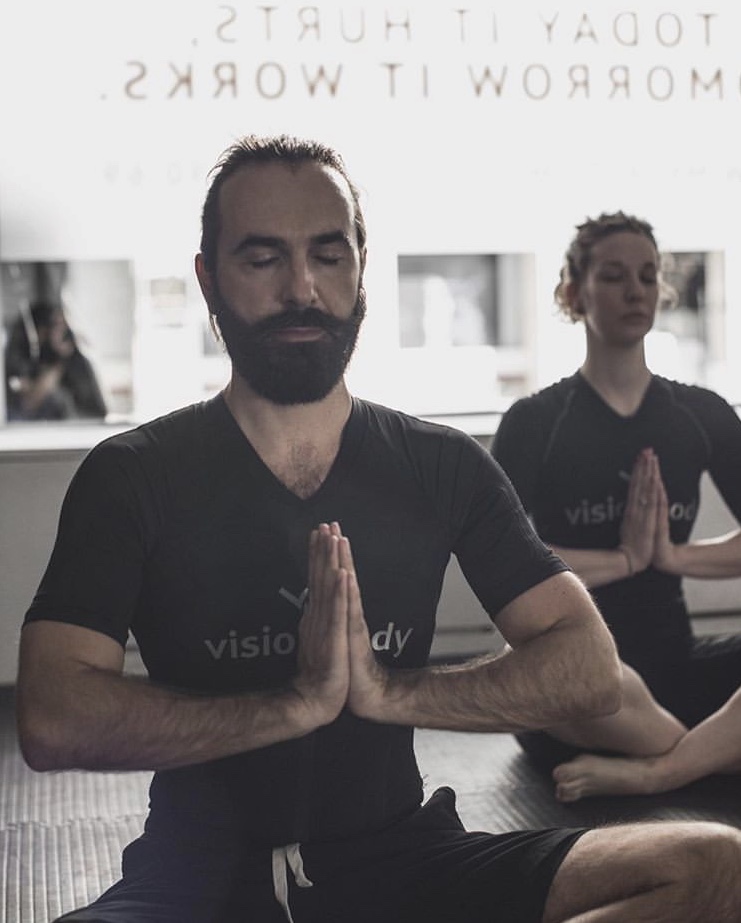 People practicing meditation in a yoga class, sitting cross-legged on the floor with hands in prayer position. A quote on the wall behind them reads: "Today it hurts, tomorrow it works," inspiring them to stay committed to their weight loss goals.