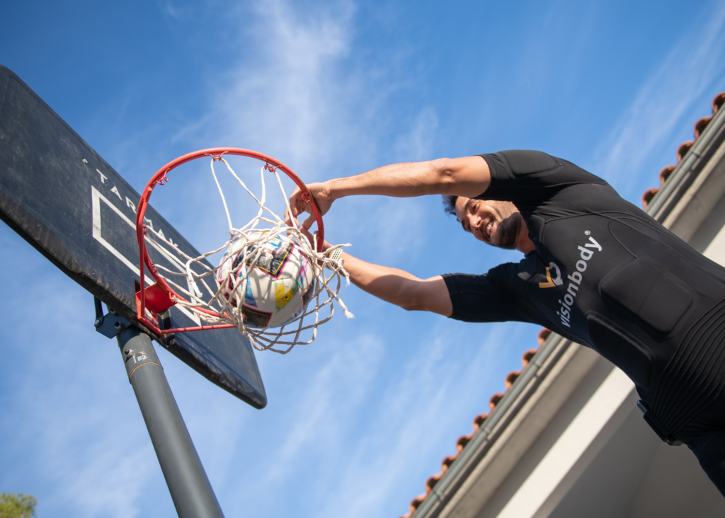 A person in a black VisionBody shirt stands beside a basketball hoop, either placing or retrieving a basketball.